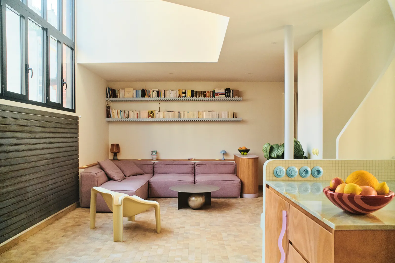 Dans le salon un canap modulable Cosima lilas et un fauteuil jaune pâle Lounge F300 font face à la cuisine. Au sol des...